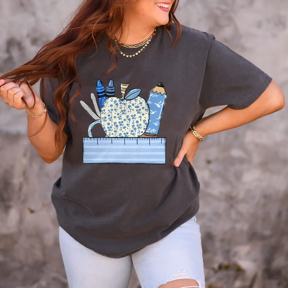 An Apple With A Blue Flower Pattern Surrounded By Stationery, Including Blue Crayons, Pencils, Scissors And Paper Clips, And A Light Blue Ruler At The Bottom, With A Casual T-Shirt.