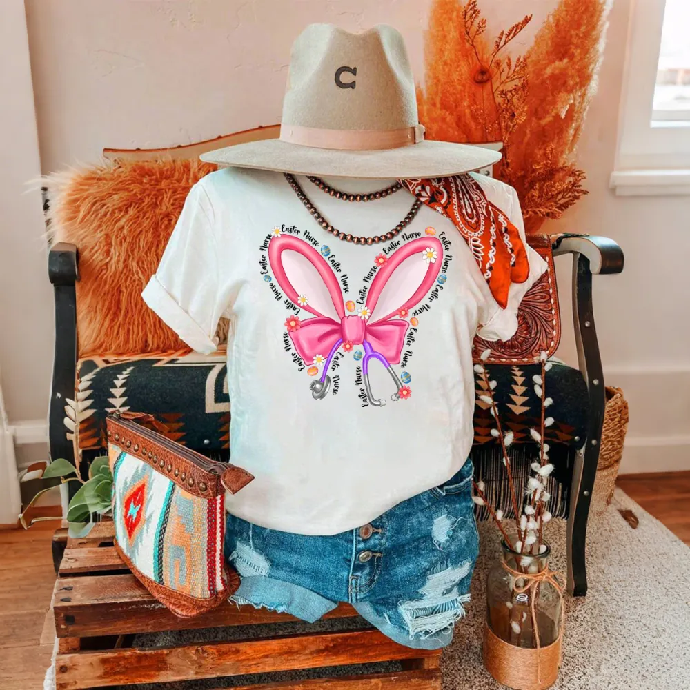 Repeating “Easter Nurse” Multiple Times, The Slogan Is Wrapped Around Pink Cartoon Bunny Ears And Bows, A Stethoscope, And Is Dotted With Small Flowers And Gems, And A Casual T-Shirt.